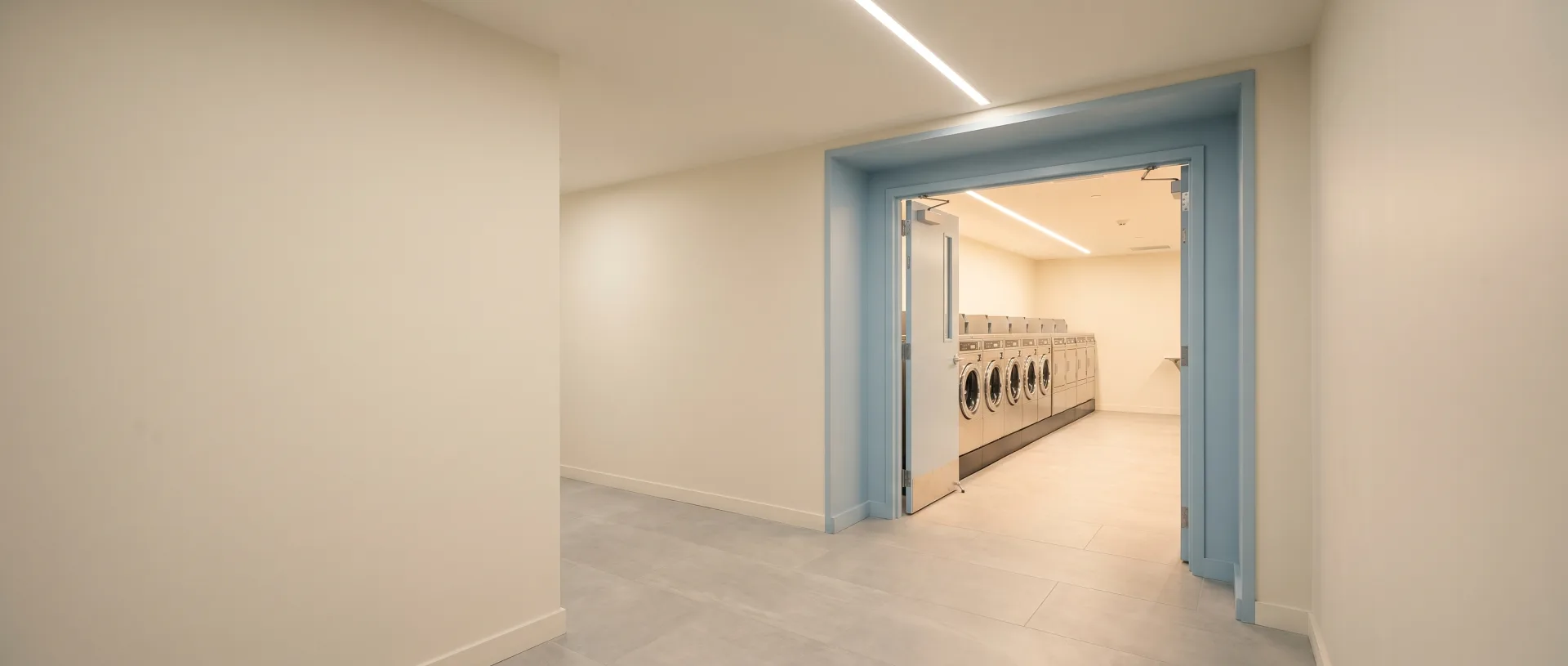 A clean apartment corridor with warm light spilling from a wide doorway into a large finished laundry amenity facility.