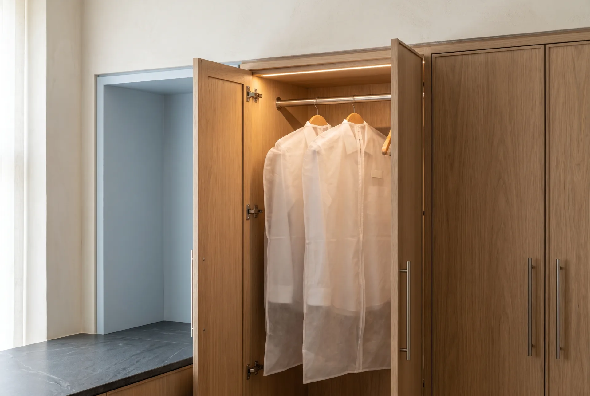 An open white oak garment locker holding two clear garment bags with pressed shirts on wooden hangers, beside closed locker doors with brushed nickel pulls.