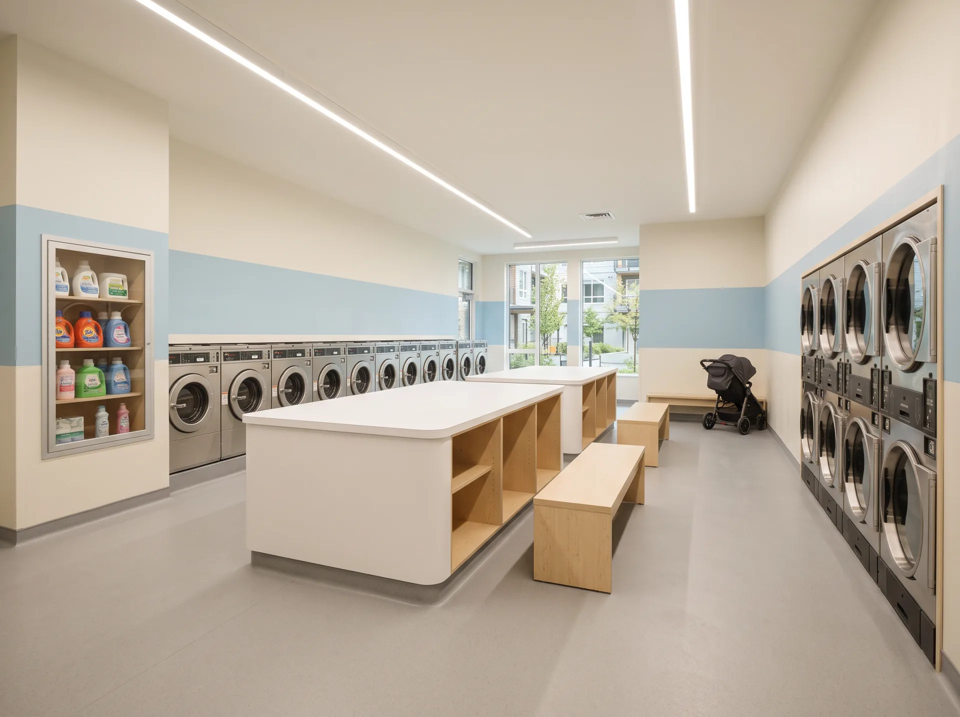 Large bright family-building laundry facility with eighteen washers, twelve stacked dryers, light gray resilient flooring, a long laminate folding counter and a powder-blue accent band.